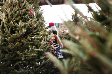 Lächelnde Frau, die ihr Smartphone in der Hand hält und inmitten von Weihnachtsbäumen steht - CAVF17009