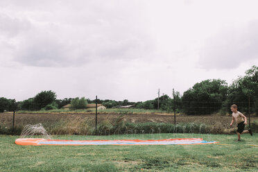 Shirtless boy playing on water slide at yard against sky - CAVF17239