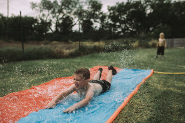 Boy sliding on water slide at yard with brother standing in background - CAVF17241