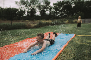 Junge rutscht auf einer Wasserrutsche im Garten mit seinem Bruder im Hintergrund - CAVF17241