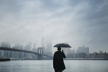 Rückansicht eines Mannes, der einen Regenschirm hält, während er auf eine Stadtansicht vor einem bewölkten Himmel blickt - CAVF17252