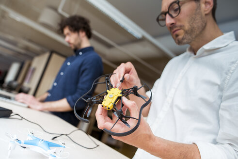 Techniker, der eine Drohne untersucht, während ein Kollege im Büro einen Laptop benutzt - CAVF17383