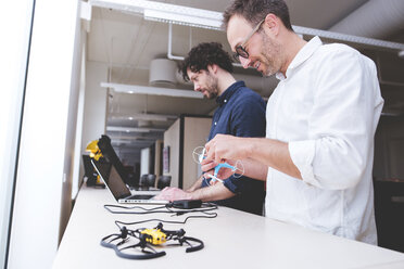 Techniker, der eine Drohne untersucht, während ein Kollege im Büro einen Laptop benutzt - CAVF17396