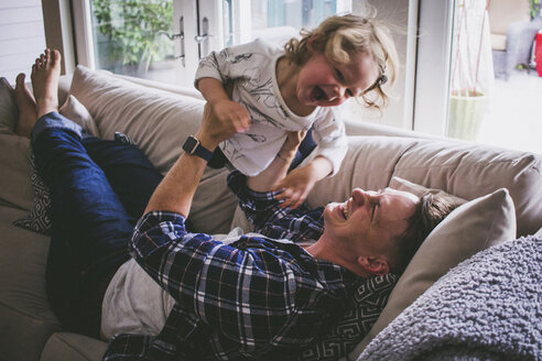 Glücklicher Vater hebt seine Tochter hoch, während er zu Hause auf dem Sofa liegt - CAVF17441