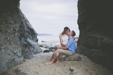 Romantisches Paar, das sich küsst, während es auf einem Felsen am Strand sitzt - CAVF17449