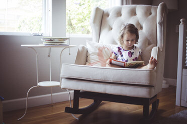Mädchen liest Bilderbuch, während sie zu Hause auf einem Schaukelstuhl sitzt - CAVF17453