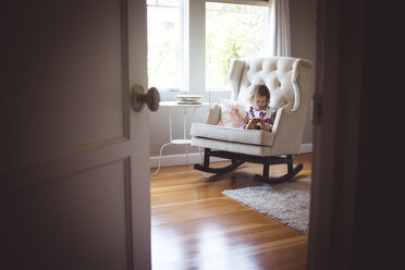Mädchen liest Bilderbuch, während sie auf einem Schaukelstuhl zu Hause sitzt, gesehen durch die Türöffnung - CAVF17454