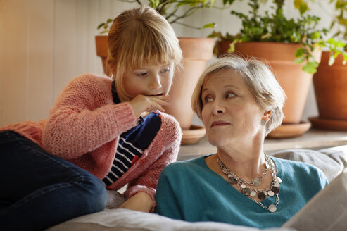 Großmutter betrachtet ihre Enkelin beim Lesen einer Zeitschrift, während sie auf dem Sofa sitzt - CAVF17617