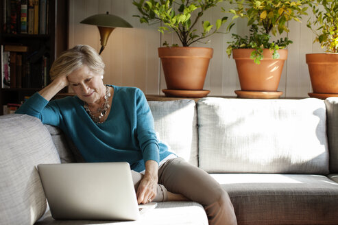 Frau mit Hand im Haar, die einen Laptop benutzt, während sie zu Hause auf dem Sofa sitzt - CAVF17623