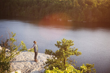 High angle view of young man standing with arms outstretched on rock overlooking lake - CAVF17642