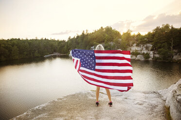 Rückansicht einer Frau mit amerikanischer Flagge auf einem Felsen mit Blick auf den See - CAVF17647