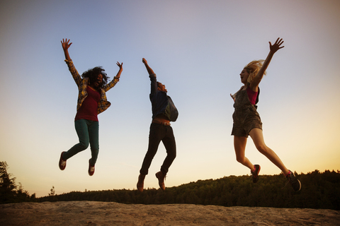 Freunde springen in der Abenddämmerung auf einem Felsen gegen den klaren Himmel, lizenzfreies Stockfoto