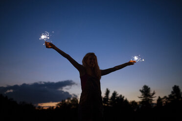 Cheerful woman with arms outstretched holding sparklers against sky at night - CAVF17667