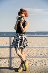 Young woman photographing through camera while standing by railing at beach - CAVF17671