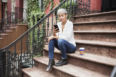 Woman using mobile phone while sitting on steps with coffee cup - CAVF17689