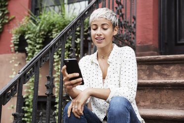 Happy woman using mobile phone while sitting on steps - CAVF17693