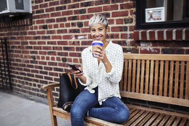 Porträt einer Frau, die auf einer Bank sitzend ein Mobiltelefon und eine Kaffeetasse hält - CAVF17699