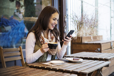 Smiling woman holding coffee cup while using mobile phone at cafe - CAVF17729