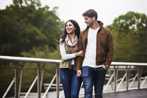 Glückliches Paar hält sich an den Händen, während es auf einer Brücke gegen den klaren Himmel läuft - CAVF17733