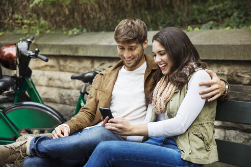 Paar benutzt Mobiltelefon, während es auf einer Parkbank sitzt - CAVF17751