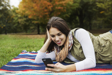 Glückliche Frau, die ein Mobiltelefon benutzt, während sie auf einer Decke im Park liegt - CAVF17766