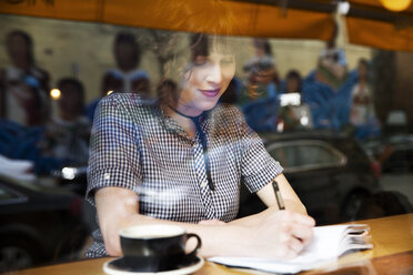 Woman writing on book while sitting in cafe seen through window - CAVF17785