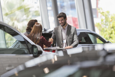 Car salesman handshaking with customers in car dealership showroom - CAIF19996