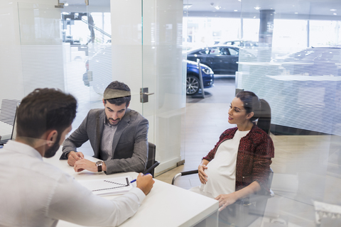 Ein Autoverkäufer erklärt einem schwangeren Paar im Büro eines Autohauses den finanziellen Vertragspapierkram, lizenzfreies Stockfoto