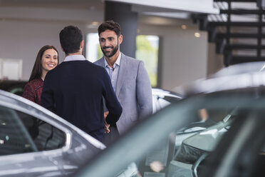 Car salesman greeting, shaking hands with couple customers in car dealership showroom - CAIF20038