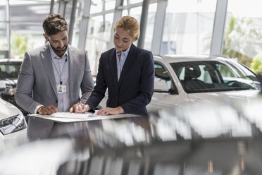 Car salesman watching female customer signing financial contract paperwork in car dealership showroom - CAIF20040