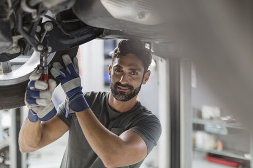 Porträt selbstbewusster männlicher Automechaniker bei der Arbeit unter einem Auto in einer Autowerkstatt - CAIF20044
