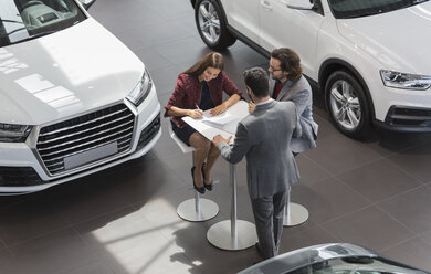 Car salesman watching couple customers signing financial contract paperwork in car dealership showroom - CAIF20047