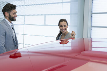 Ein lächelnder Autoverkäufer zeigt einer Kundin im Ausstellungsraum eines Autohauses ein neues rotes Auto - CAIF20077