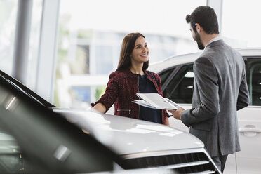 Car saleswoman showing brochure to male customer in car dealership showroom - CAIF20089