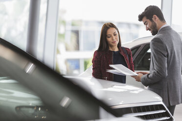 Car saleswoman showing brochure to male customer in car dealership showroom - CAIF20098