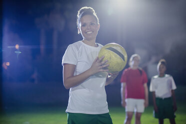 Portrait smiling, confident young female soccer player holding soccer ball on field at night - CAIF20107