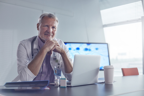 Porträt eines selbstbewussten männlichen Mediziners, der in einem Krankenhaus am Laptop arbeitet, lizenzfreies Stockfoto