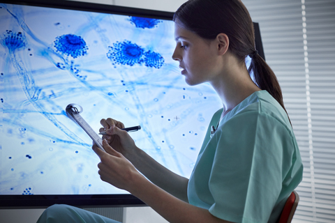 Krankenschwester mit Klemmbrett, die einen vergrößerten Objektträger auf einem Computermonitor betrachtet, lizenzfreies Stockfoto