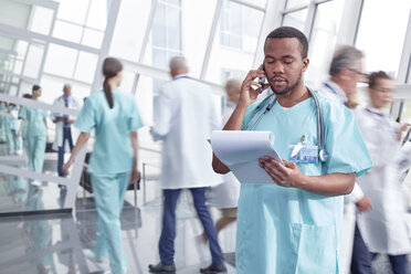 Männlicher Chirurg mit Klemmbrett, der in der Lobby eines Krankenhauses mit einem Handy telefoniert - CAIF20179