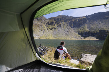 Male athlete sitting by tent against lake - CAVF17853