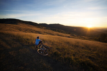 Männlicher Sportler mit Fahrrad auf einer Wiese sitzend gegen den Himmel bei Sonnenuntergang - CAVF17877