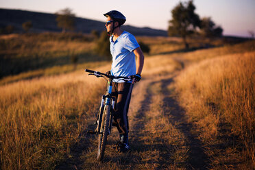 Full length of male cyclist standing with bicycle on dirt road field - CAVF17878