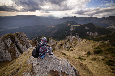 Rear view of male backpacker sitting on mountain against cloudy sky - CAVF17896