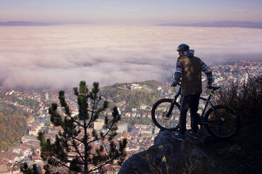 Rear view of cyclist looking at cityscape while standing on cliff - CAVF17903