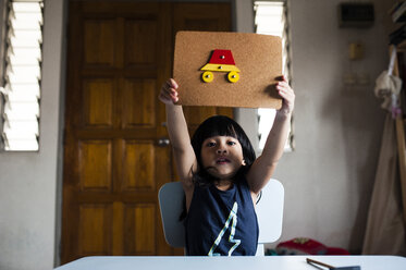 Porträt eines niedlichen Jungen, der am Tisch sitzend seine auf Pappe gefertigten Kunstwerke zeigt - CAVF17961