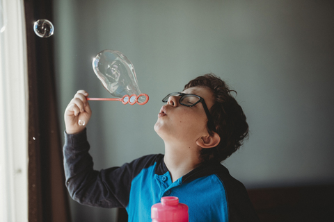 Boy wearing eyeglasses blowing bubbles at home stock photo