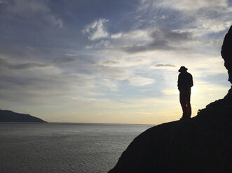 Silhouette eines Mannes, der bei Sonnenuntergang auf einem felsigen Berg am Meer steht - CAVF18058