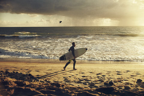 Mann mit Surfbrett beim Strandspaziergang bei Sonnenuntergang, lizenzfreies Stockfoto