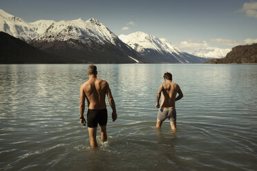 Rear view shirtless male friends walking into Kenai lake against snowcapped mountains - CAVF18085