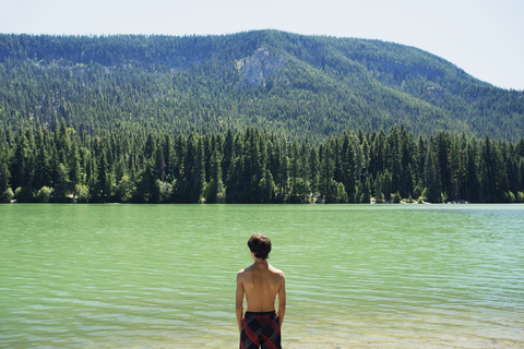 Rückansicht eines Jungen am See vor einem Berg an einem sonnigen Tag, lizenzfreies Stockfoto